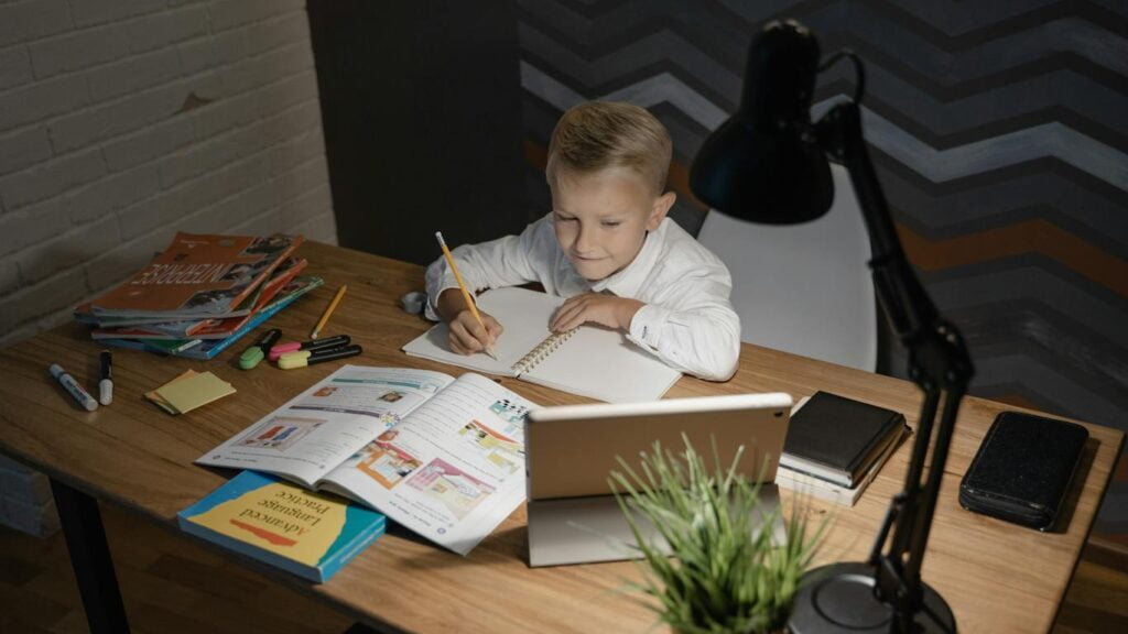 A young boy sits at a desk, engrossed in his laptop and book, on a study table designed for students